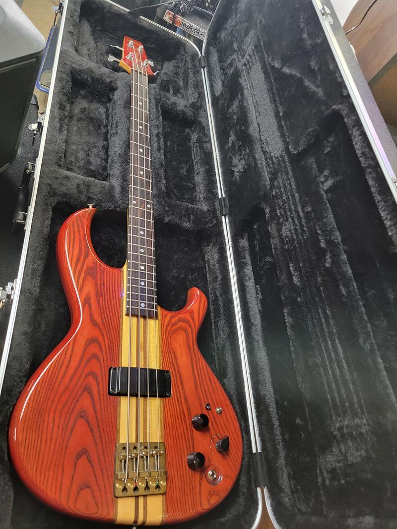 Natural-finish four-string bass with through-neck stripe and brass bridge in an open hardshell case